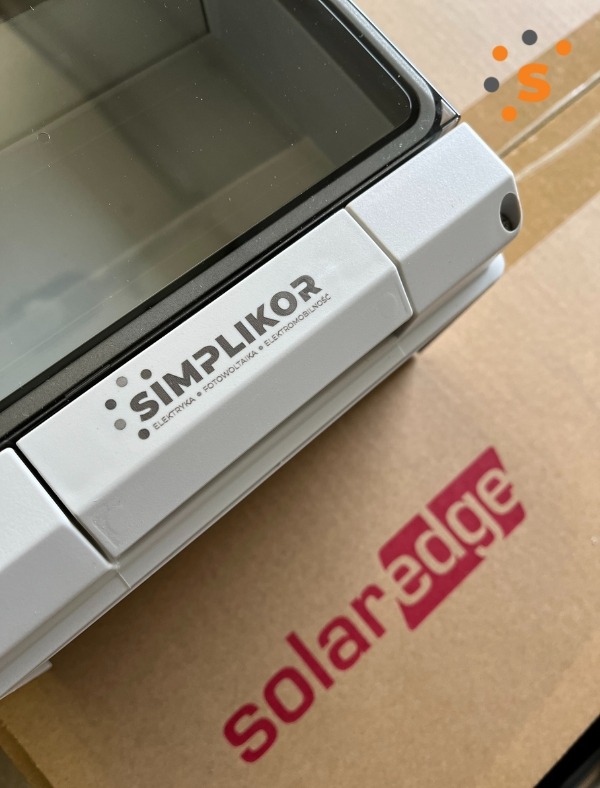 Biała obudowa z przezroczystym okienkiem, oznaczona logo 'SIMPLIKOR' z informacją 'ELEKTRYKA FOTOWOLTAIKA ELEKTROMOBILNOŚĆ', leżąca na kartonie z czerwonym logo 'solaredge'.