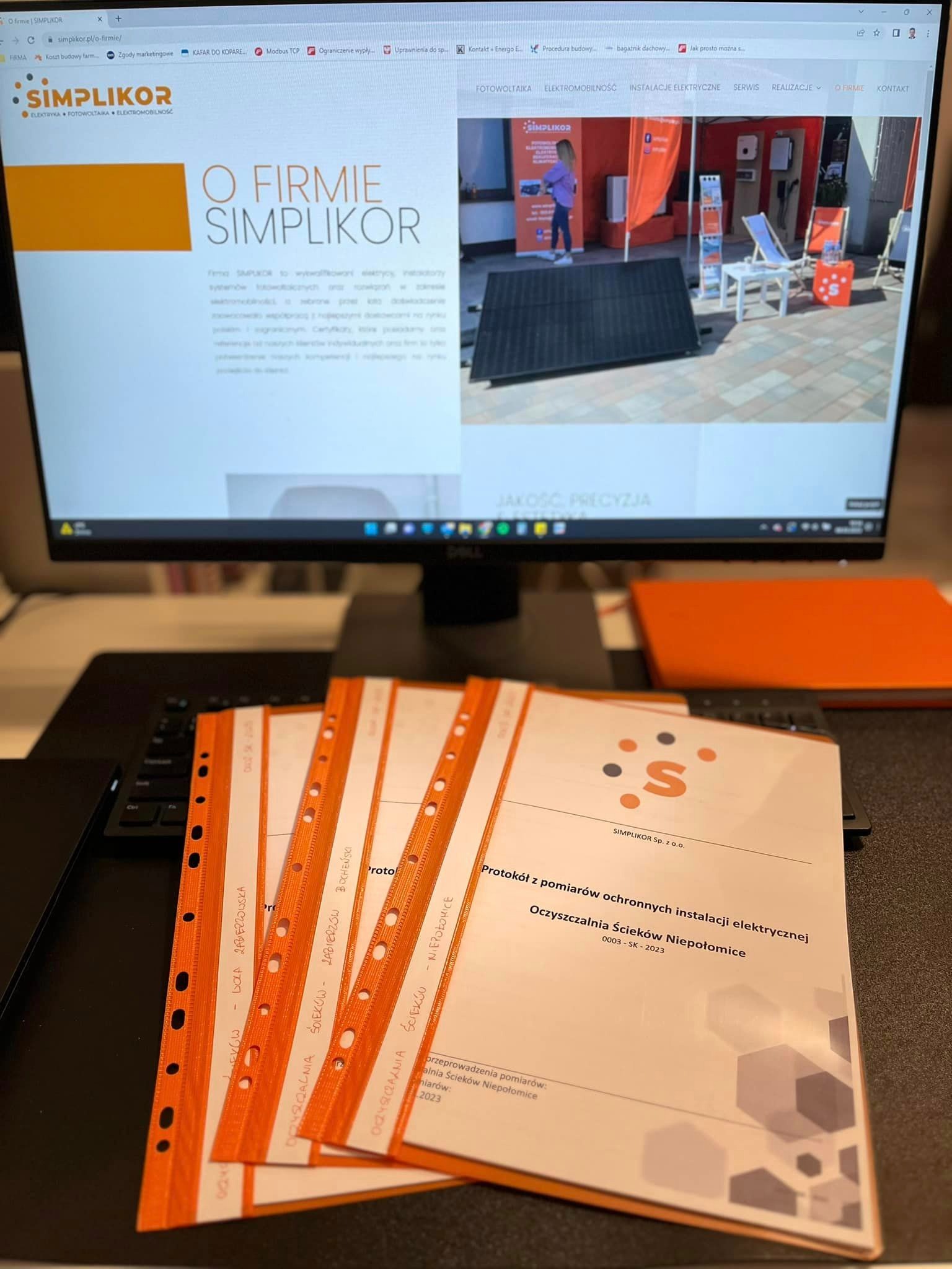 Ekran monitora wyświetlający stronę internetową firmy Simplikor z ofertą fotowoltaiki i instalacji elektrycznych, poniżej leżą dokumenty w pomarańczowych segregatorach z protokołami pomiarów...