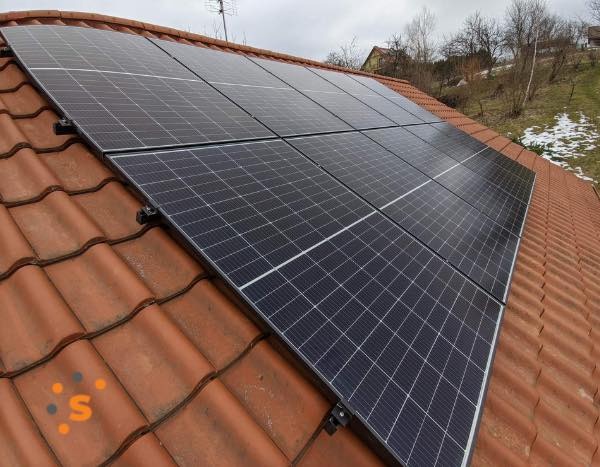 Panele słoneczne zamontowane na dachu pokrytym czerwoną dachówką ceramiczną, widoczne mocowania paneli, w tle fragment krajobrazu z drzewami i resztkami śniegu.