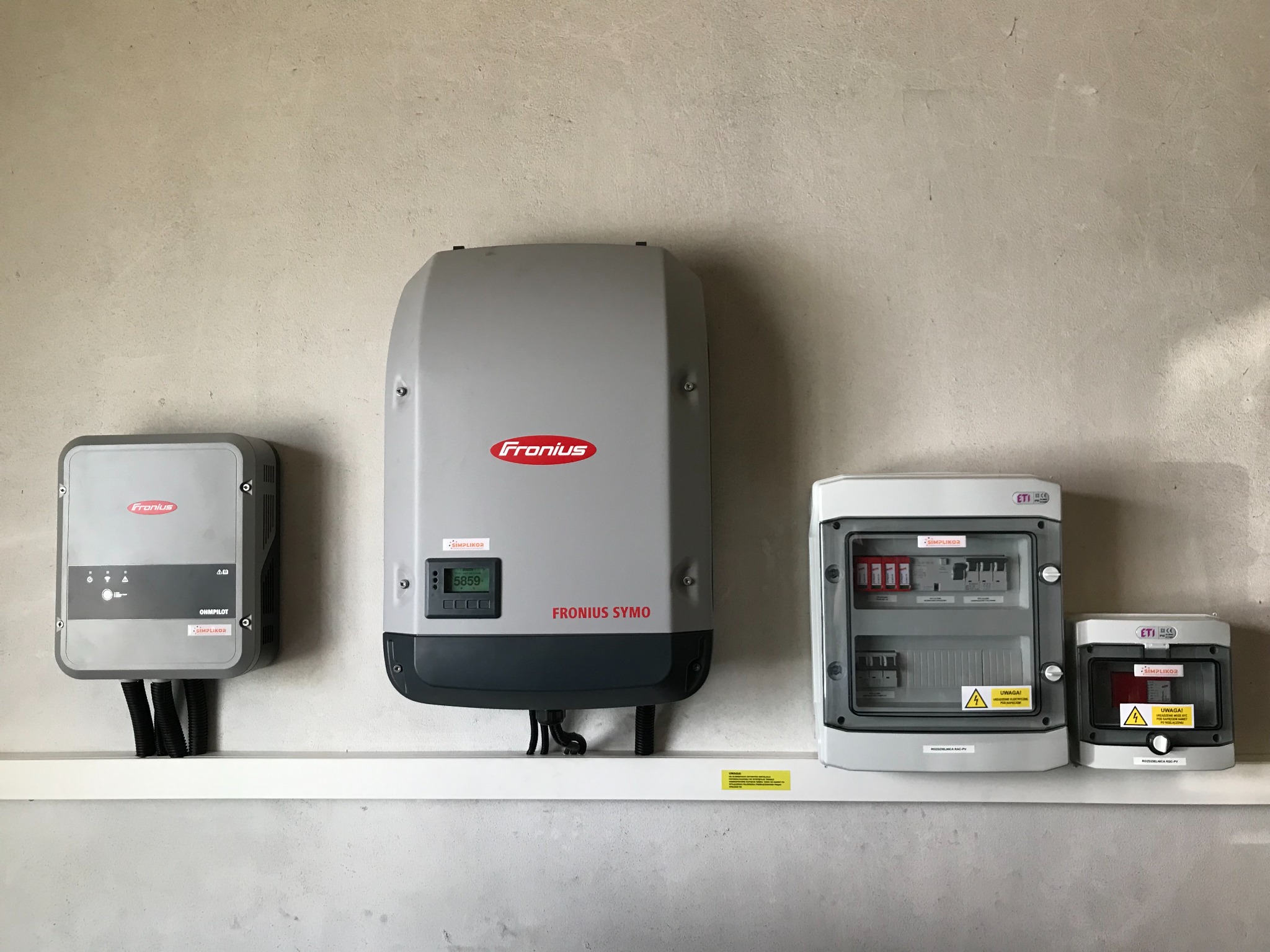 Szare inwertery Fronius i rozdzielnice ETI zamontowane na ścianie nad białą półką, widoczne kable w peszlach, tabliczki ostrzegawcze i wyświetlacz z wartością 5859.