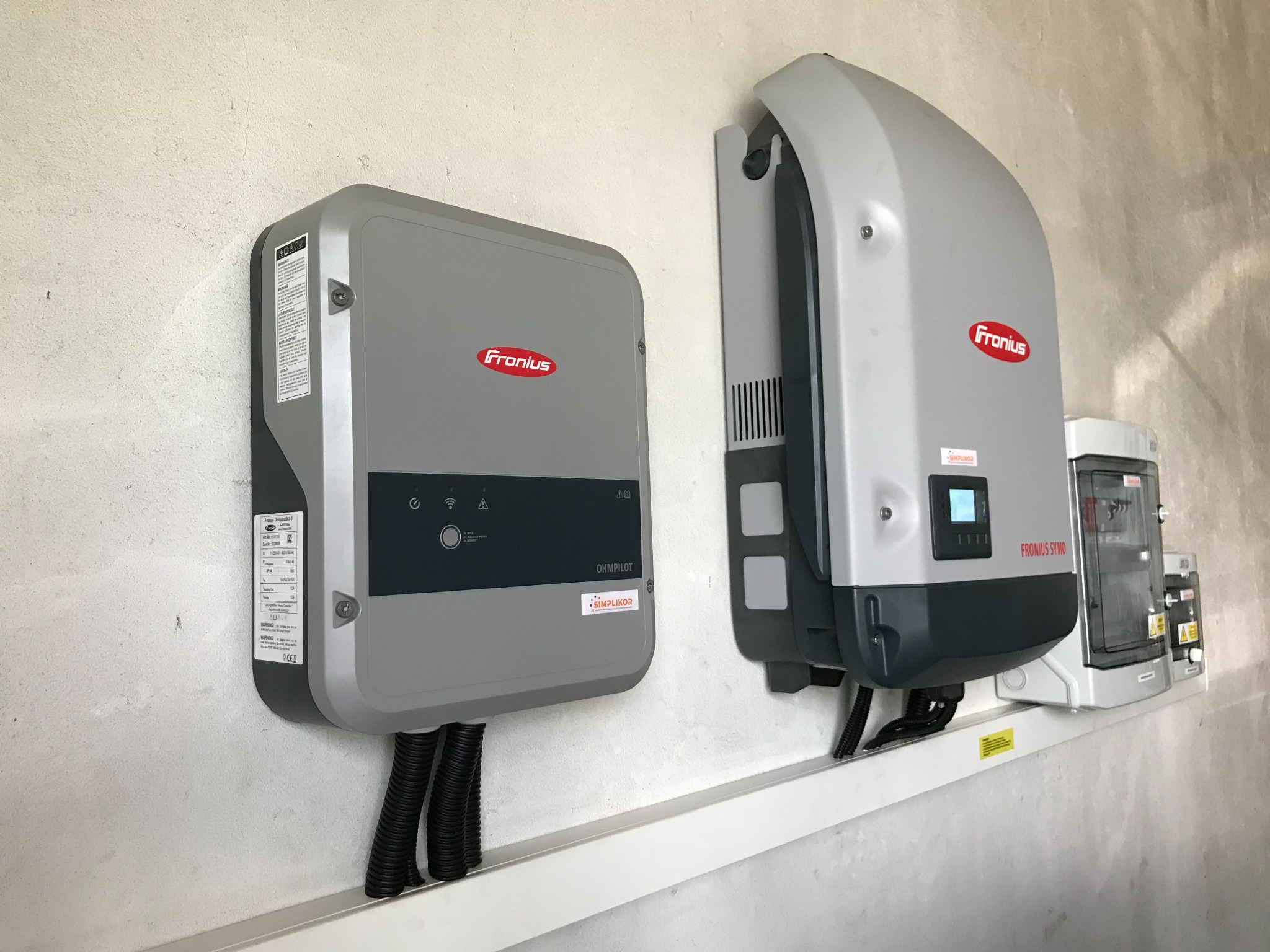 Dwa szare inwertery marki Fronius zamontowane na ścianie, z widocznymi przewodami w peszlach i dodatkową skrzynką elektryczną obok, na tle surowej ściany.