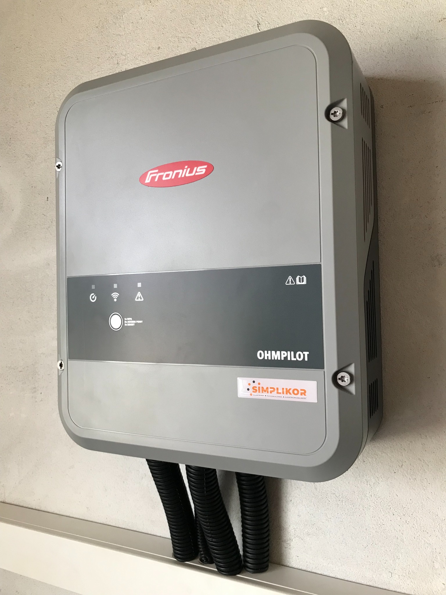 Szary regulator Fronius OHMPILOT zamontowany na ścianie, z podłączonymi trzema czarnymi rurami karbowanymi, widoczne logo Simplikor.