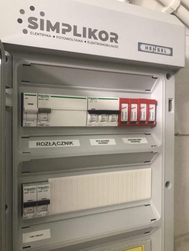 Szara skrzynka elektryczna marki Hensel z widocznymi bezpiecznikami, wyłącznikami i naklejkami informacyjnymi, w tym rozłącznik główny i wyłącznik przepięć. Na górze logo firmy Simplikor zajmującej...