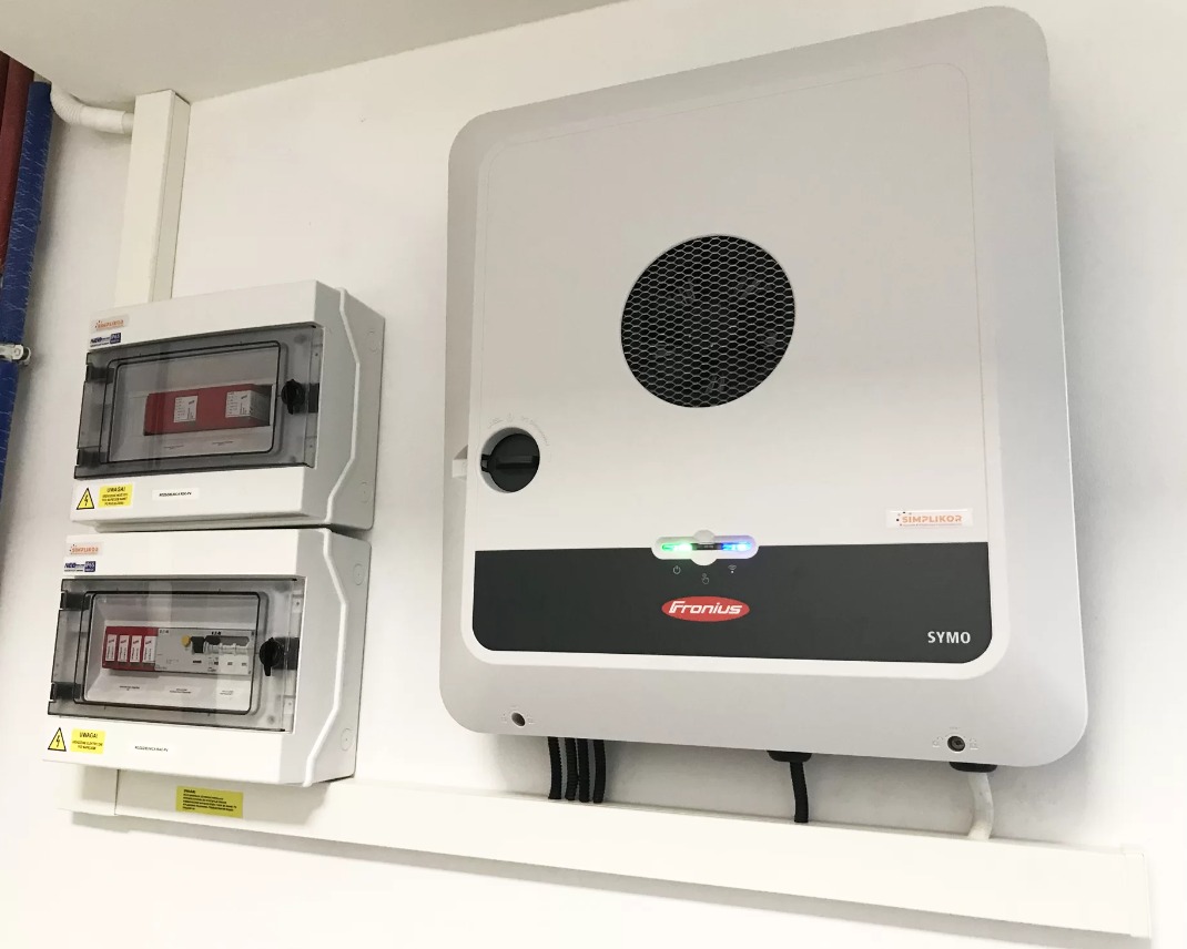 Biały inwerter Fronius SYMO zamontowany na ścianie obok dwóch skrzynek rozdzielczych z bezpiecznikami, widoczne przewody elektryczne i fragment instalacji rurowej.