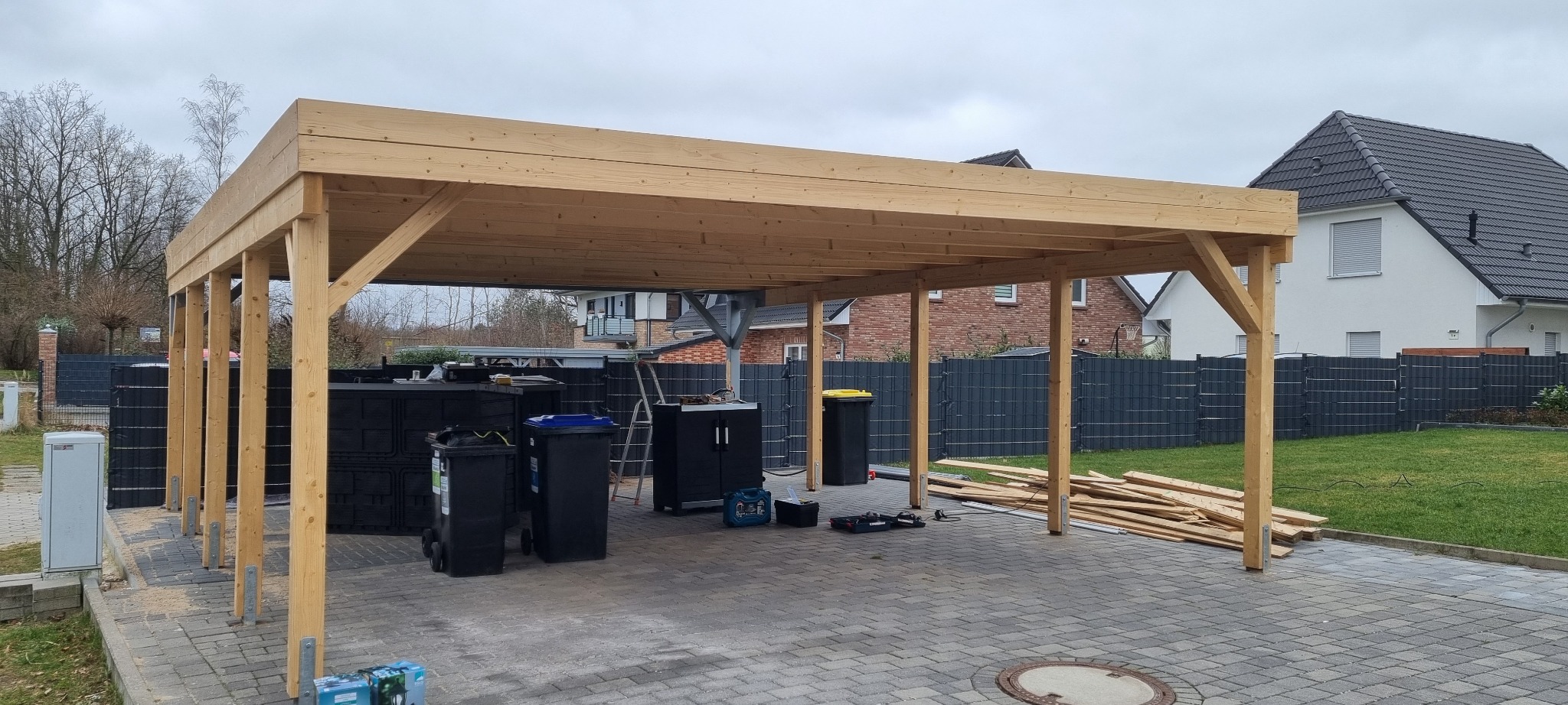 Drewniana konstrukcja z belek i słupów w trakcie budowy, widoczne ślady obróbki drewna, narzędzia i deski w tle, kostka brukowa na podłożu.