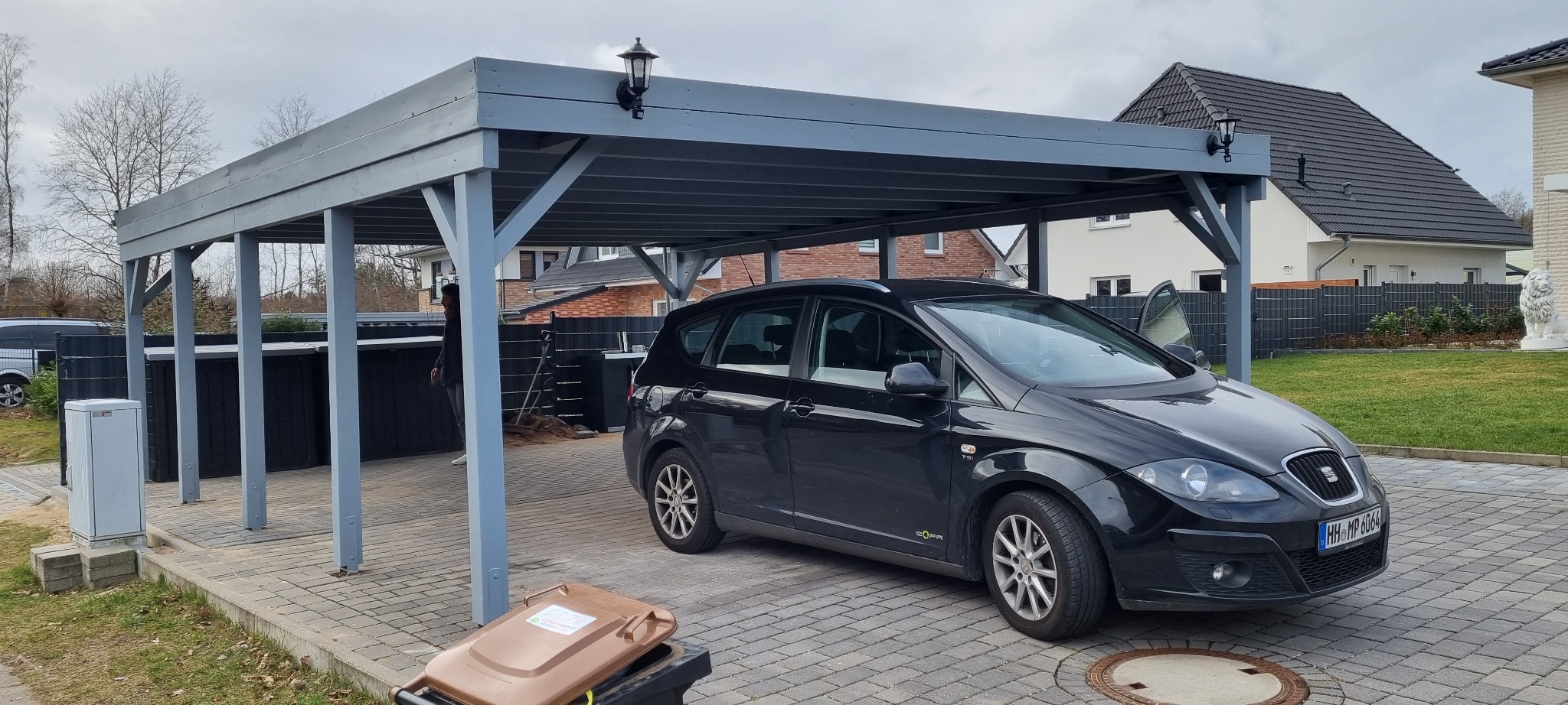 Szary drewniany carport z oświetleniem zewnętrznym, pod którym stoi czarny samochód na kostce brukowej. W tle widoczny dom z ciemnym dachem i ogrodzenie.