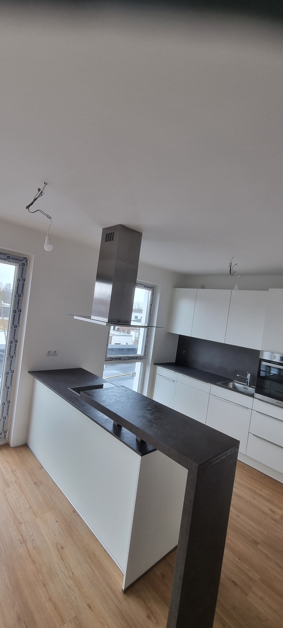 Nowoczesna kuchnia z wyspą i blatem w odcieniach szarości, białe szafki, okap kominowy ze stali nierdzewnej, surowe oświetlenie w trakcie instalacji, podłoga z paneli imitujących drewno.