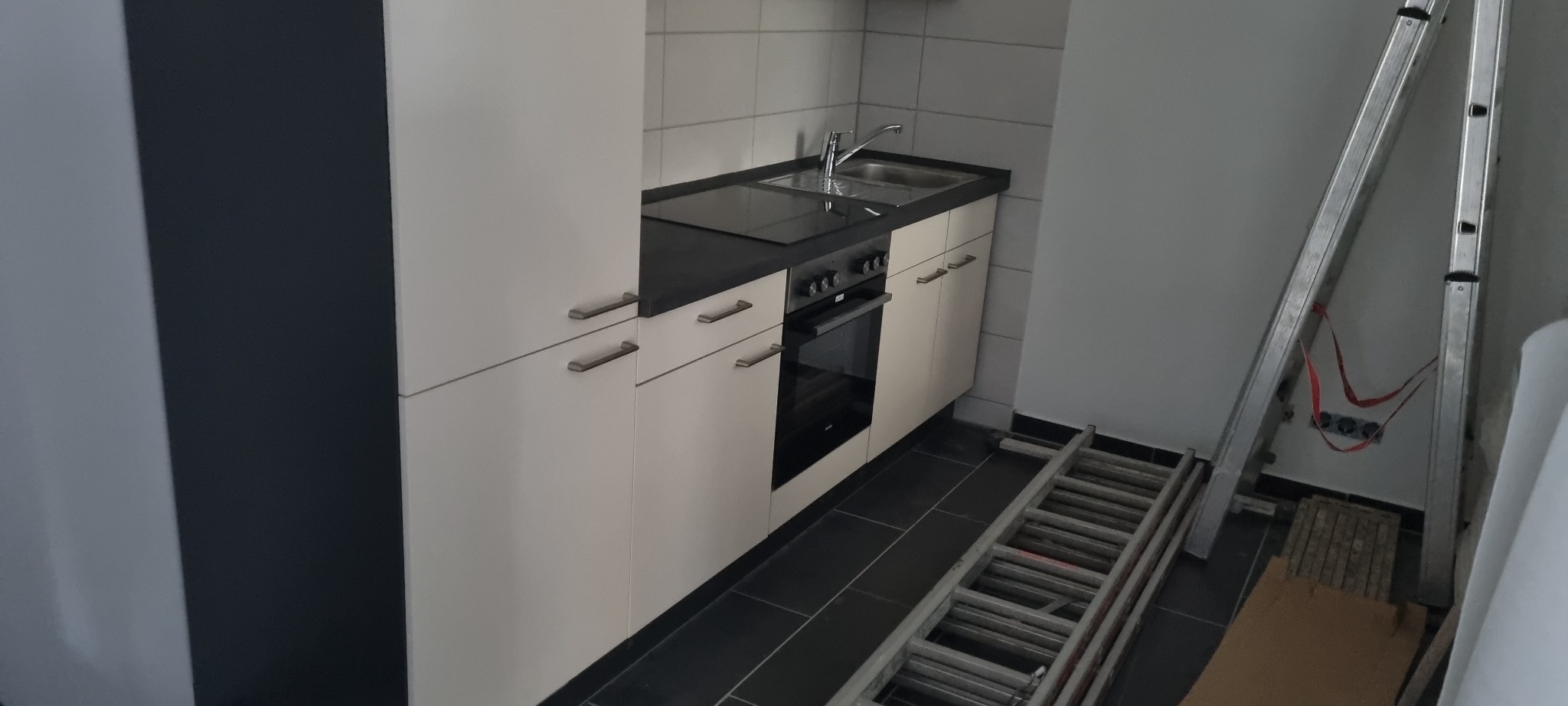 Nowoczesna, częściowo zamontowana kuchnia z białymi szafkami, czarnym blatem i płytą indukcyjną, widoczna drabina oparta o ścianę, sugerująca trwający montaż.