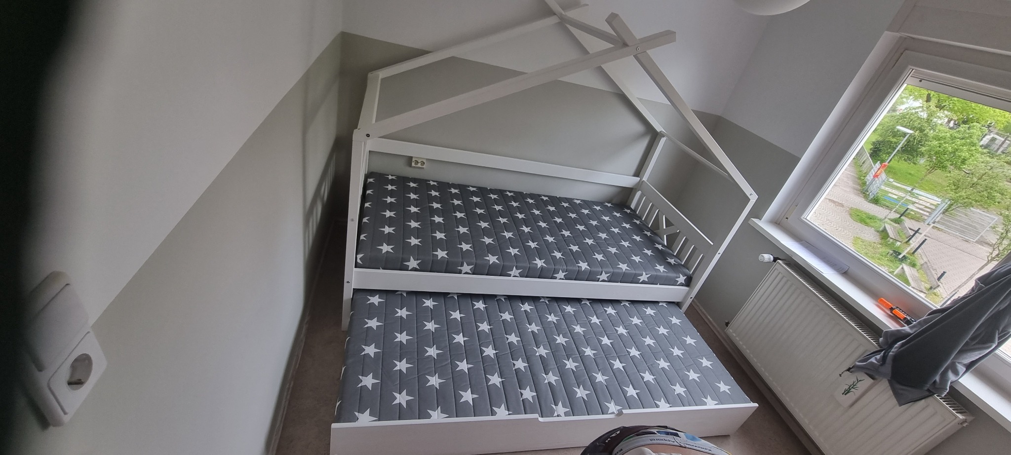 Dwuosobowe białe łóżko w kształcie domku z szarymi materacami w białe gwiazdki, ustawione w pokoju z szaro-białymi ścianami i oknem z widokiem na zewnątrz.