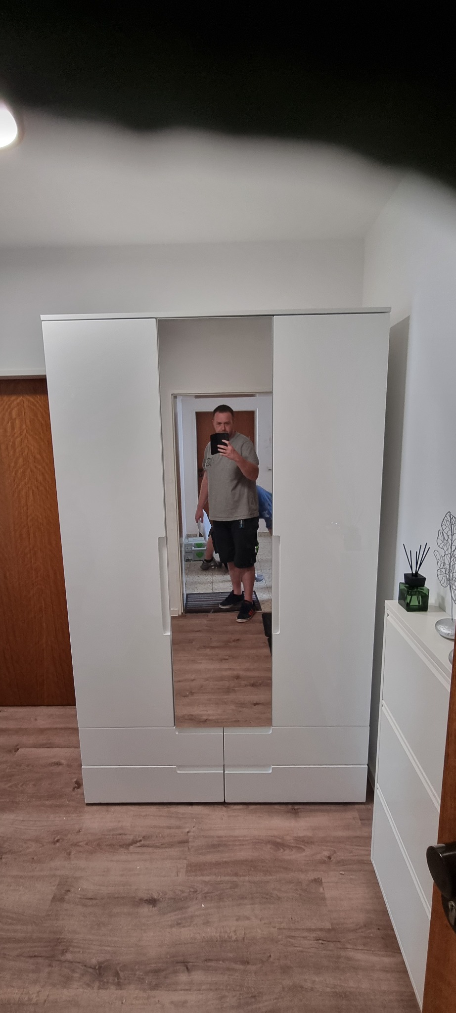 Biała, trzydrzwiowa szafa z lustrem na środkowych drzwiach, odbijająca mężczyznę robiącego zdjęcie telefonem, widoczna na tle drewnianej podłogi i białych ścian.