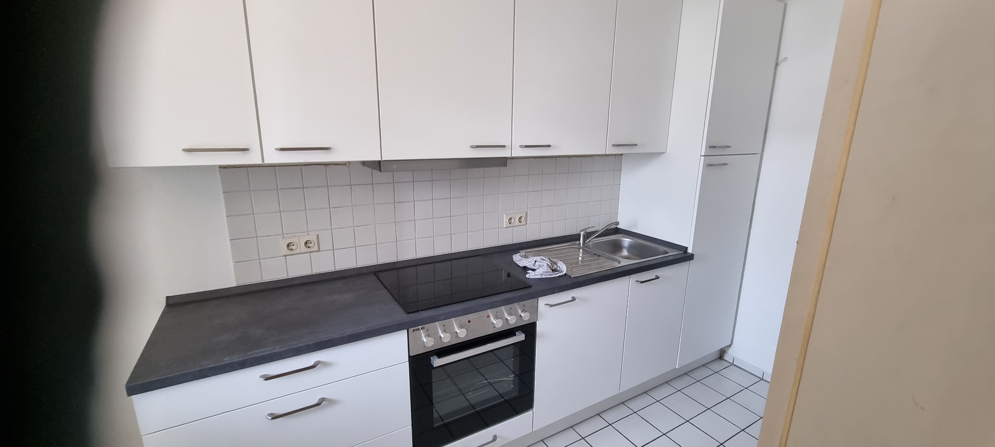 Nowoczesna, minimalistyczna kuchnia z białymi szafkami, czarnym blatem i płytkami w tle, widoczny fragment zlewu z kranem i ściereczką.