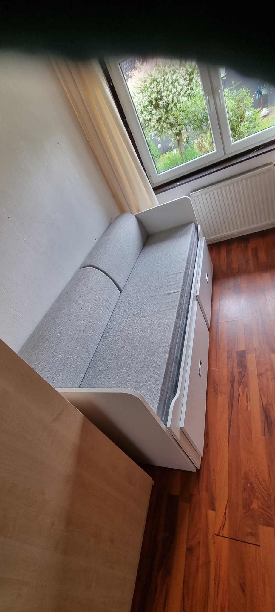 Biała antresola z szarą tapicerką, widok z góry, z widocznym oknem i grzejnikiem w tle, na podłodze panele imitujące drewno.