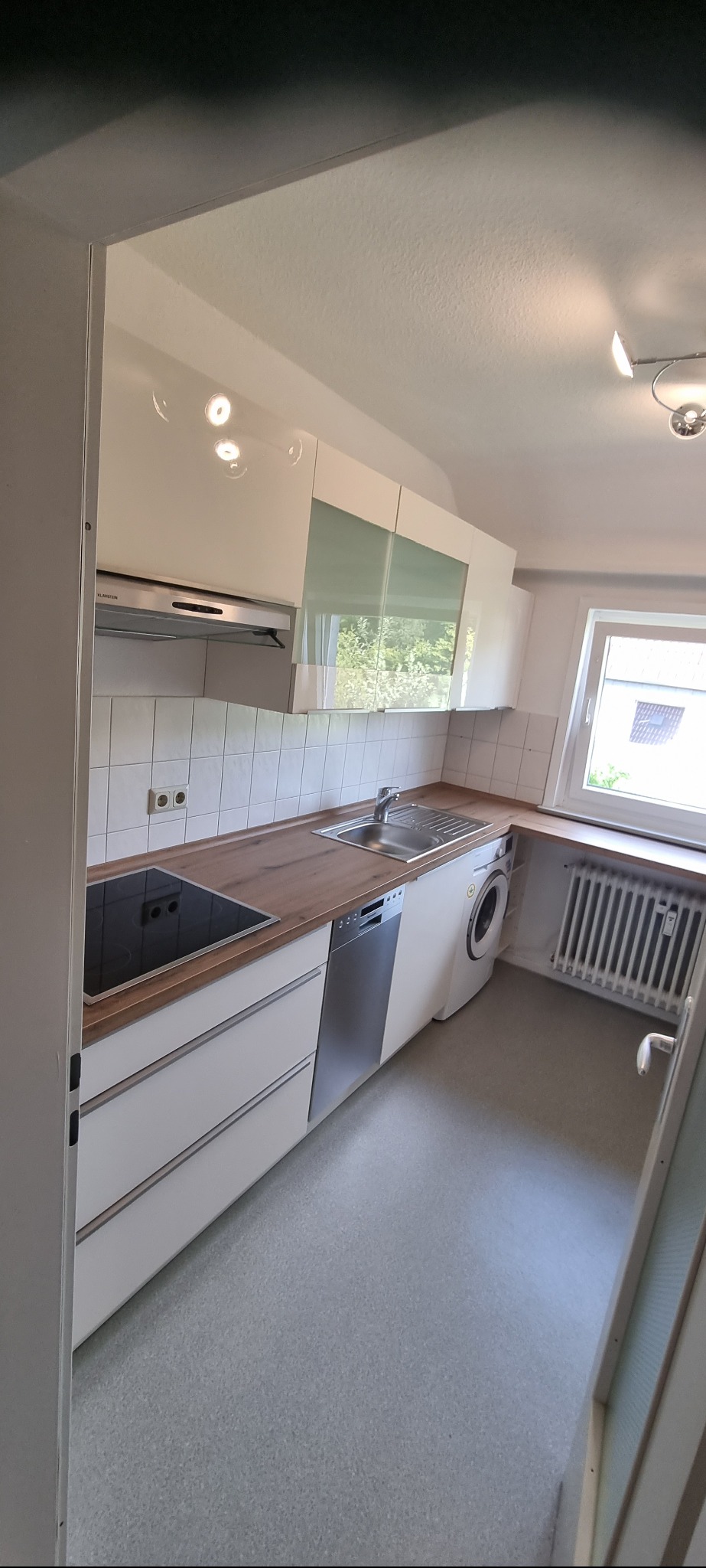 Jasna kuchnia z białymi szafkami i drewnianym blatem, z wbudowaną płytą grzewczą, zlewem, zmywarką i pralką pod blatem, widok z perspektywy wejścia do pomieszczenia.