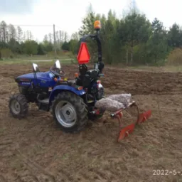Niebieski traktor z pomarańczowym światłem ostrzegawczym i workami z sadzonkami na polu przygotowanym do sadzenia, widoczne ślady opon na ziemi.