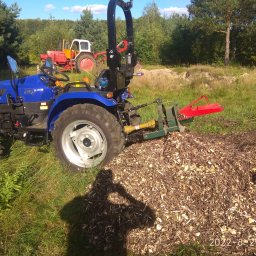 Sławomir kępa - Niebieski traktor Farmtrac z zamontowanym rozdrabniaczem gałęzi, obok sterta zrębków, w tle inny traktor i zieleń.