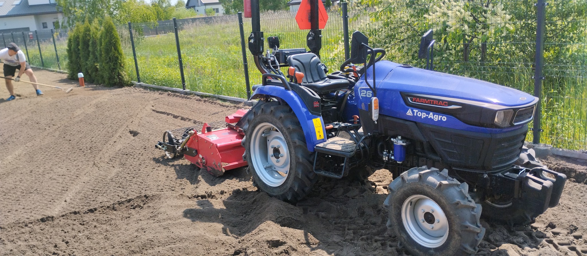 Niebieski ciągnik marki Farmtrac z czerwonym glebogryzem przygotowuje ziemię pod trawnik; w tle mężczyzna wyrównuje teren grabiami.