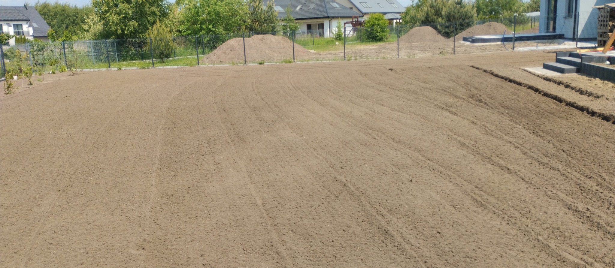 Przygotowany teren pod trawnik z wykopanymi rowkami, widoczne kopce ziemi w tle i fragment domu.