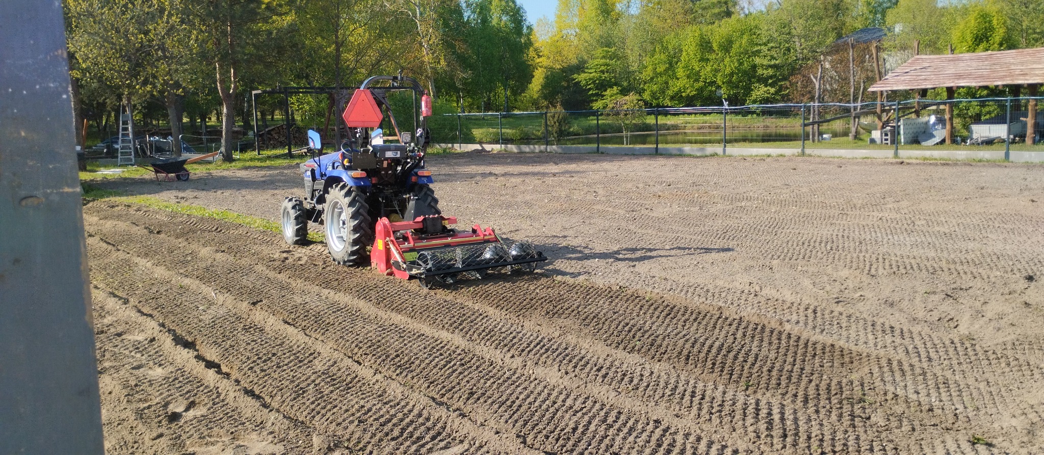 Niebieski traktor z czerwonymi elementami spulchnia ziemię na piaszczystym terenie, przygotowując grunt pod trawnik, w tle widoczny zarys budynku z drewnianym dachem i drzewa.