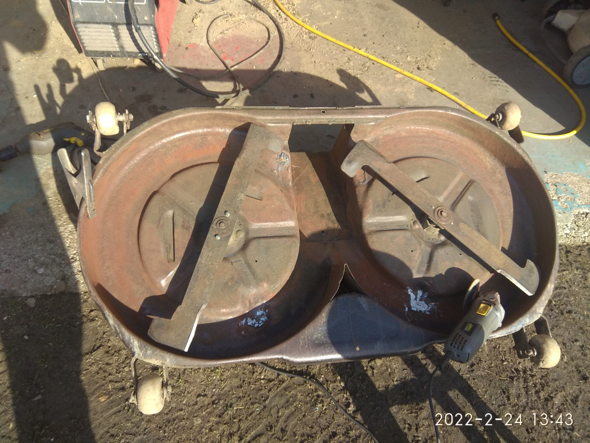 Spód kosiarki dwu-nożowej, odwrócony do góry dnem, z widocznymi ostrzami i śladami rdzy, obok szlifierka kątowa.