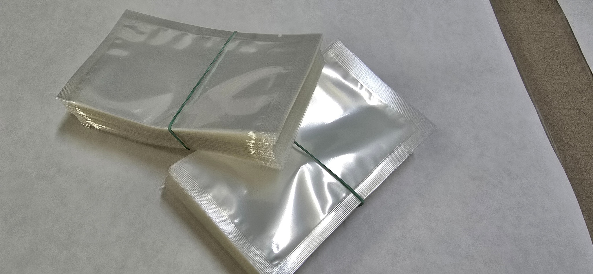 Dwa stosy przezroczystych, foliowych saszetek zgrzewanych na krawędziach, spiętych zieloną gumką recepturką, leżące na jasnym podłożu.