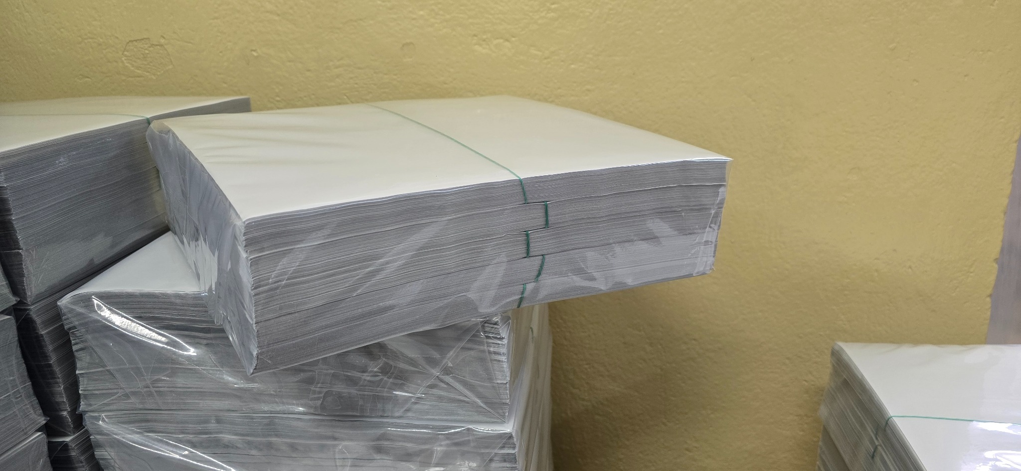 Stosy białego papieru do drukarki, owinięte folią i spięte zielonym sznurkiem, ułożone jeden na drugim na tle żółtej ściany.