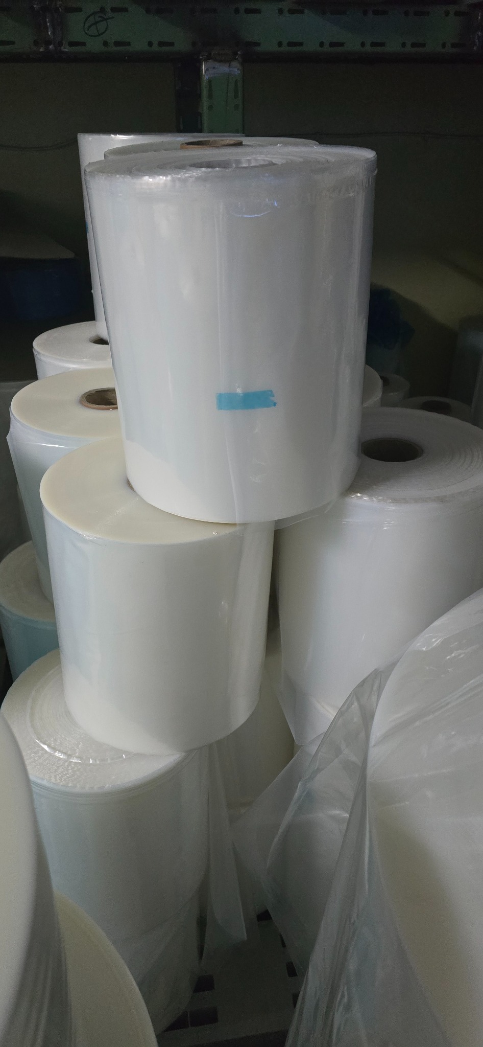 Stos rolek transparentnej folii, owiniętych w przezroczystą folię ochronną, z niebieską taśmą na jednej z nich, ułożonych na palecie w magazynie.