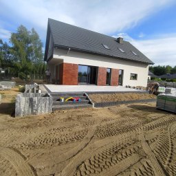 INS-BRUK-REM Stanisław Kluba Usługi brukarskie, ogólnobudowlane - Nowoczesny dom z tarasem w trakcie budowy. Widoczne ułożone schody z kostki brukowej, palety z materiałem i ślady opon na ziemi. Częściowo ułożona kostka tarasowa.