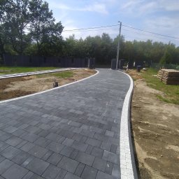 INS-BRUK-REM Stanisław Kluba Usługi brukarskie, ogólnobudowlane - Czarno-biała kostka brukowa tworząca zakrzywiony podjazd, obrzeża z jasnej kostki. W tle widoczne drzewa i elementy ogrodzenia. Słoneczny dzień.