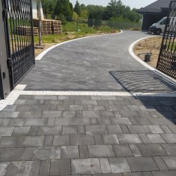 INS-BRUK-REM Stanisław Kluba Usługi brukarskie, ogólnobudowlane - Ułożona, ciemnoszara kostka brukowa z białym obramowaniem tworząca ścieżkę w ogrodzie. Widoczne metalowe ogrodzenie i budynek w tle.