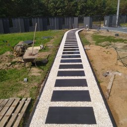 INS-BRUK-REM Stanisław Kluba Usługi brukarskie, ogólnobudowlane - Ścieżka z czarnych płyt i białego żwiru prowadząca przez trawnik, w tle ogrodzenie i drzewa. Widoczne narzędzia brukarskie.