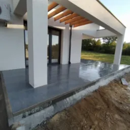 Nowoczesny taras z szarych, błyszczących płytek ceramicznych, zadaszony drewnianą pergolą, widok na ogród, klimatyzator na ścianie budynku.