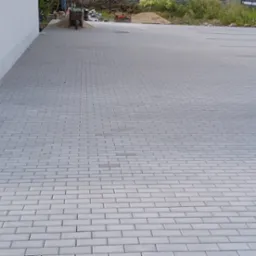 Nowa, szara kostka brukowa ułożona na parkingu, z widocznymi liniami wyznaczającymi miejsca postojowe i budynkiem w tle, częściowo widoczna ubijarka w lewym dolnym rogu.