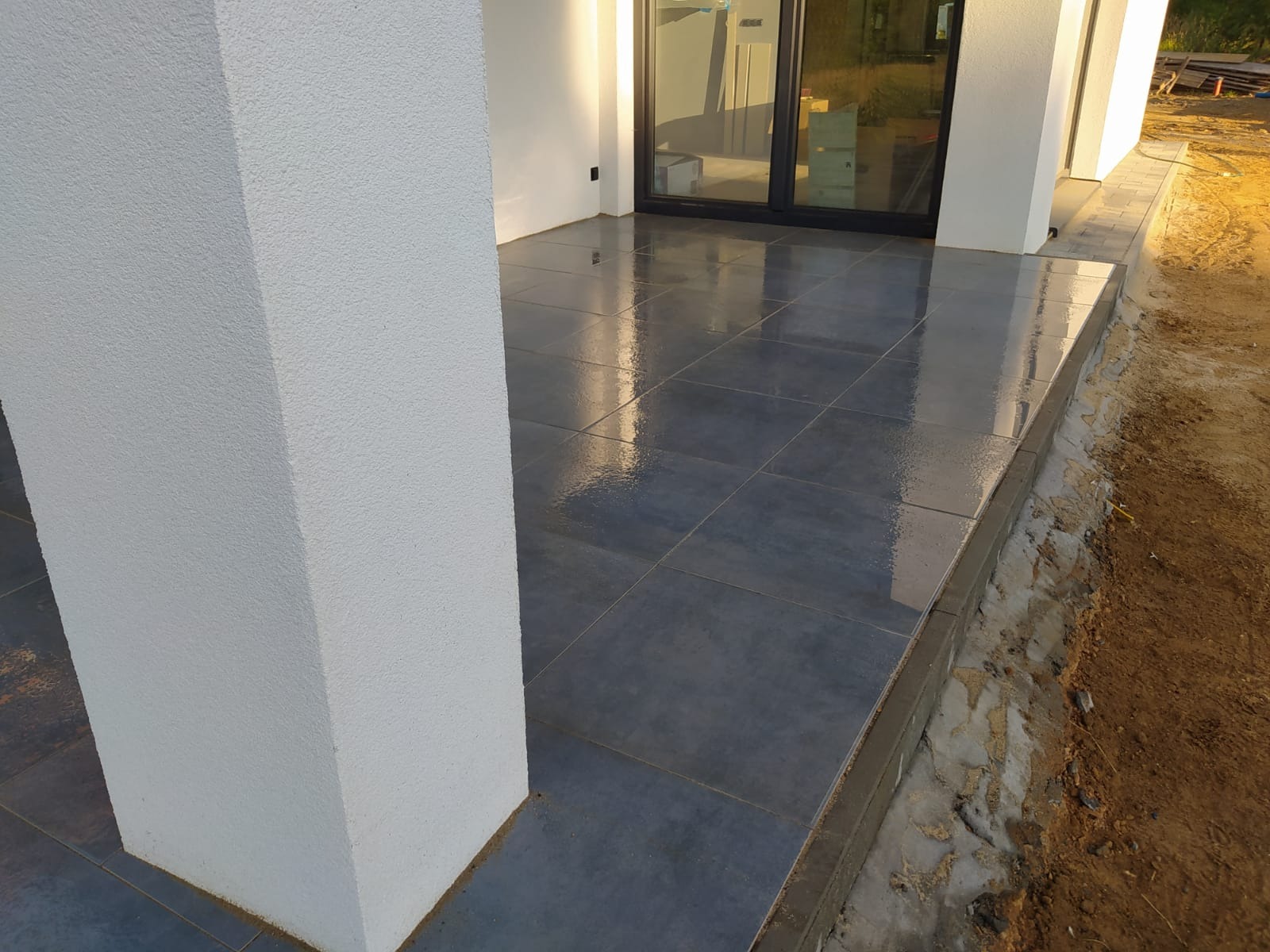 Błyszczące, ciemnoszare płytki na zewnątrz domu, ułożone na tarasie, widoczne białe kolumny i okno przesuwne, świeżo zakończone prace wykończeniowe przy krawędzi tarasu.