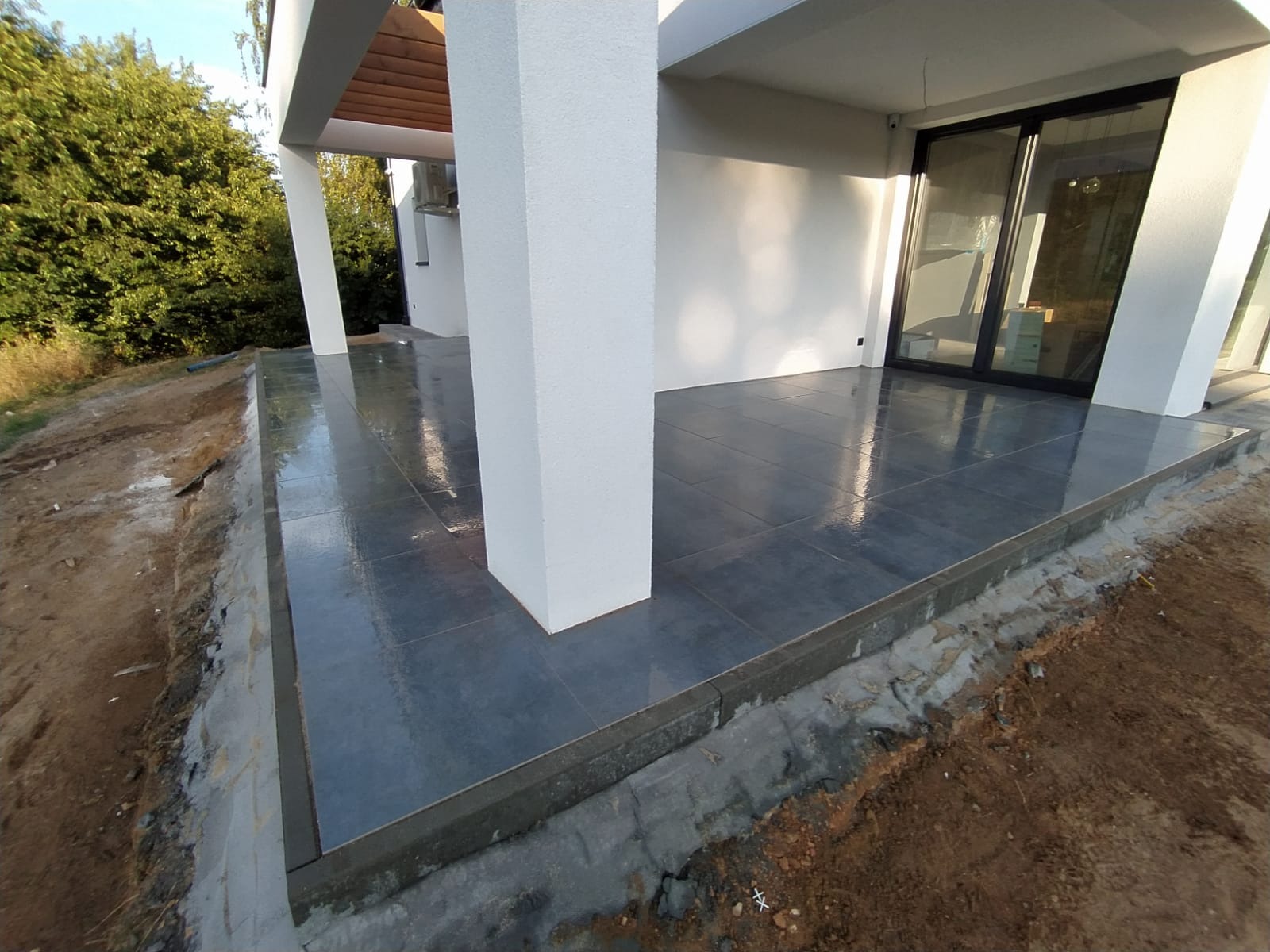 Nowoczesny taras z szarych płytek ceramicznych, widoczne białe filary, surowy brzeg tarasu z ziemią w tle.