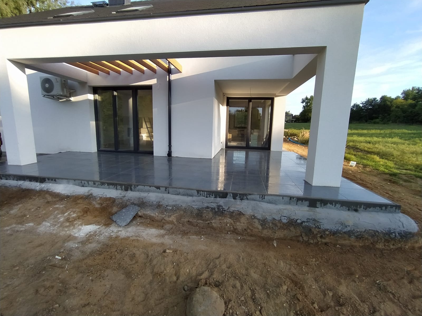 Szare płytki na tarasie nowoczesnego domu, widoczne niedokończone prace ziemne wokół, fragment niezabudowanej płytki leży na ziemi.