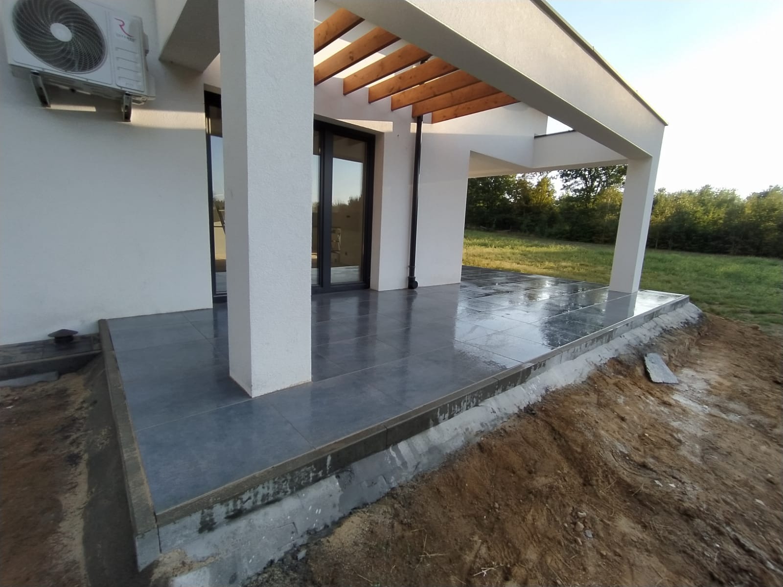 Nowoczesny taras z szarych, błyszczących płytek ceramicznych, zadaszony drewnianą pergolą, widok na ogród, klimatyzator na ścianie budynku.