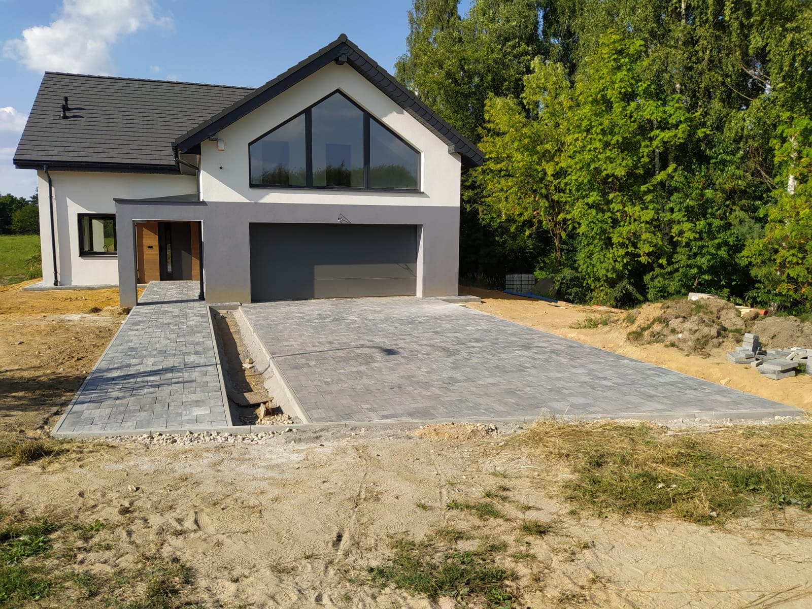 Nowoczesny dom z szarą kostką brukową na podjeździe i chodniku, widoczne niedokończone obrzeża i przygotowane materiały budowlane.