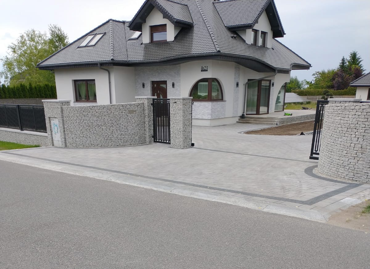 Nowoczesny dom z szarą kostką brukową na podjeździe, obrzeża z ciemniejszej kostki, ogrodzenie z kamienia i metalu.