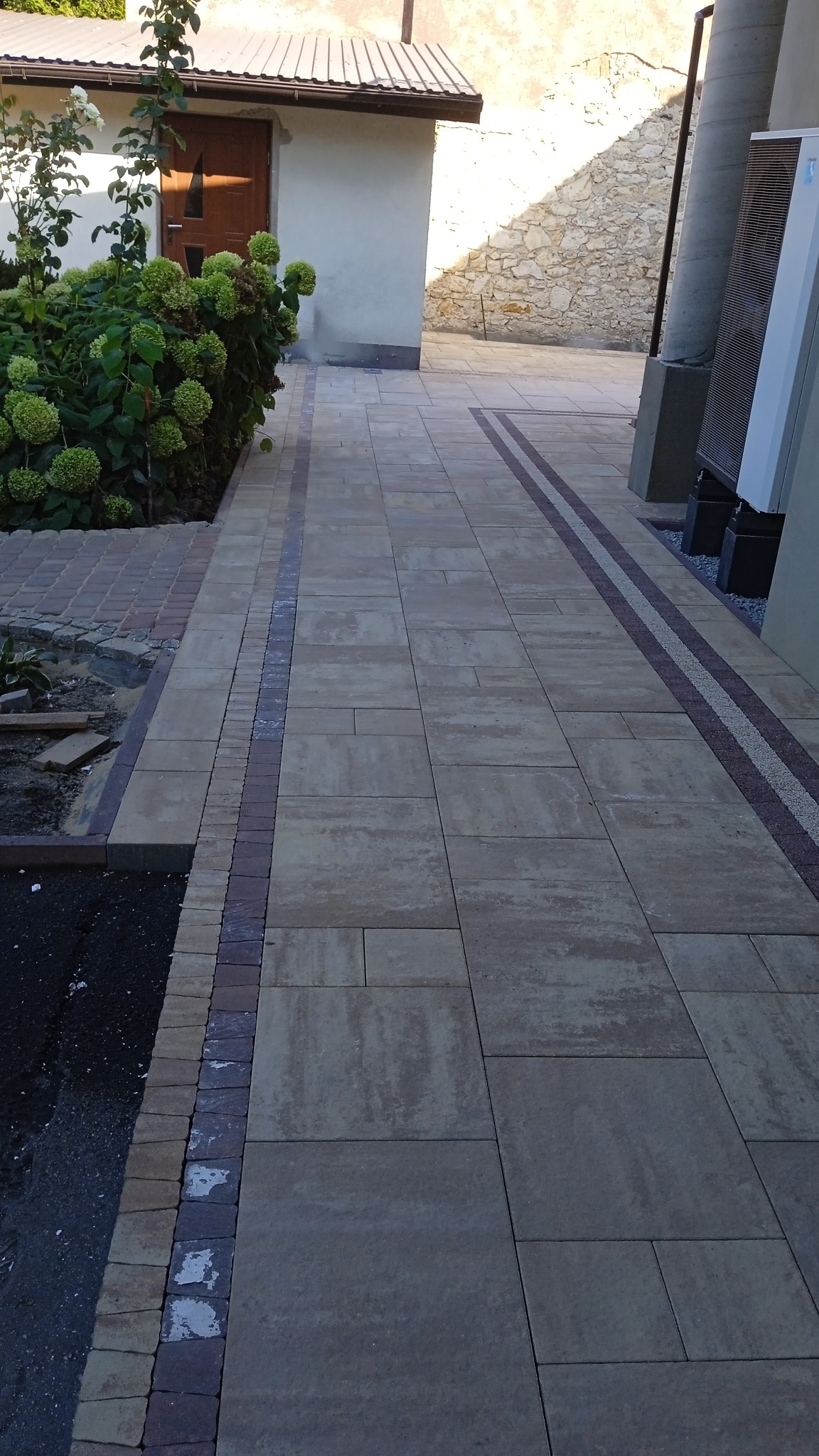 Ułożona z beżowych płyt kostka brukowa z bordowymi wstawkami, obrzeża z kostki w odcieniach brązu, widok na budynek gospodarczy i fragment ogrodu z hortensjami.