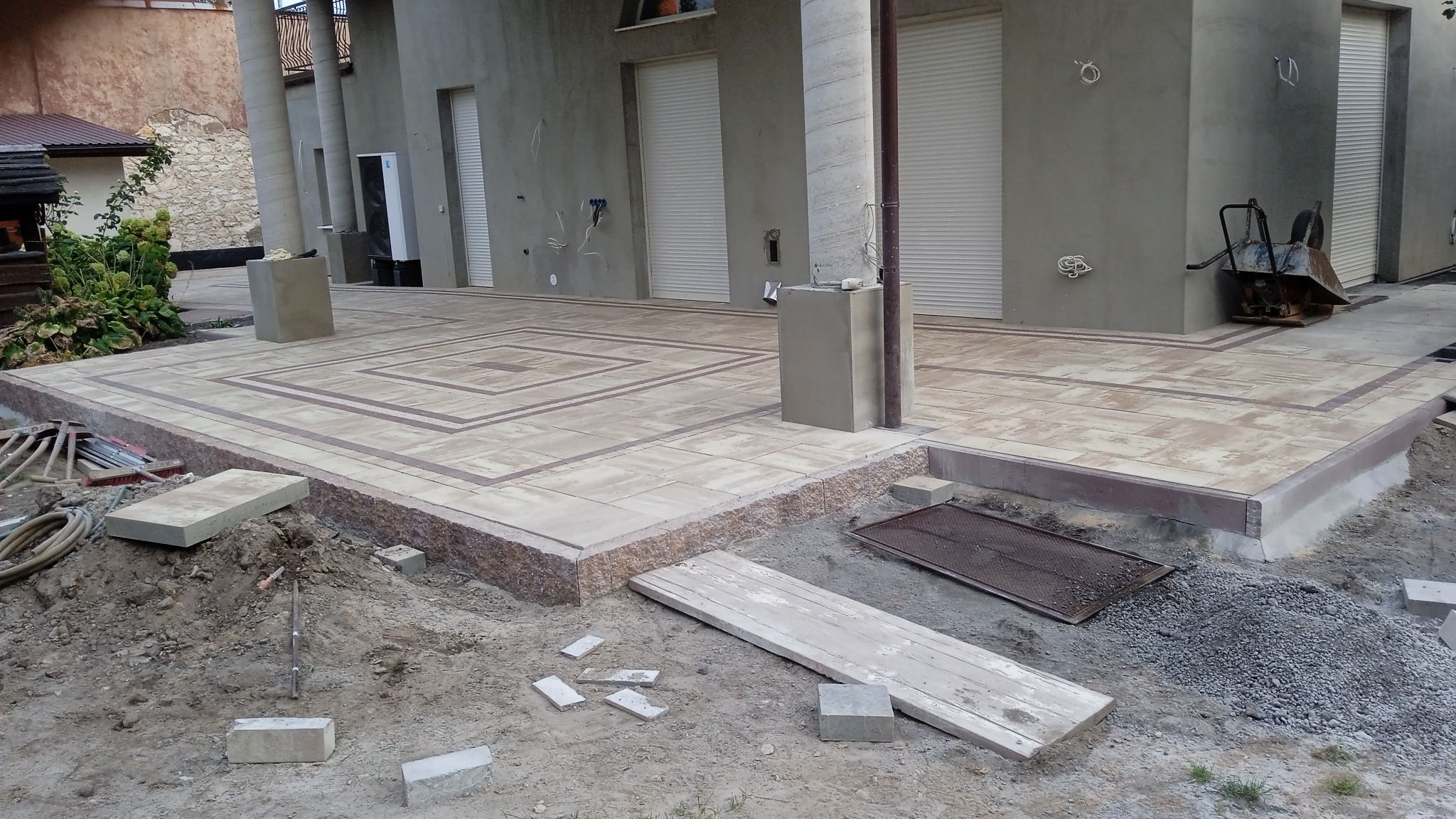 Fragment podwórka z częściowo ułożoną kostką brukową w geometryczny wzór, widoczne narzędzia i materiały budowlane, tło stanowi budynek z kolumnami.