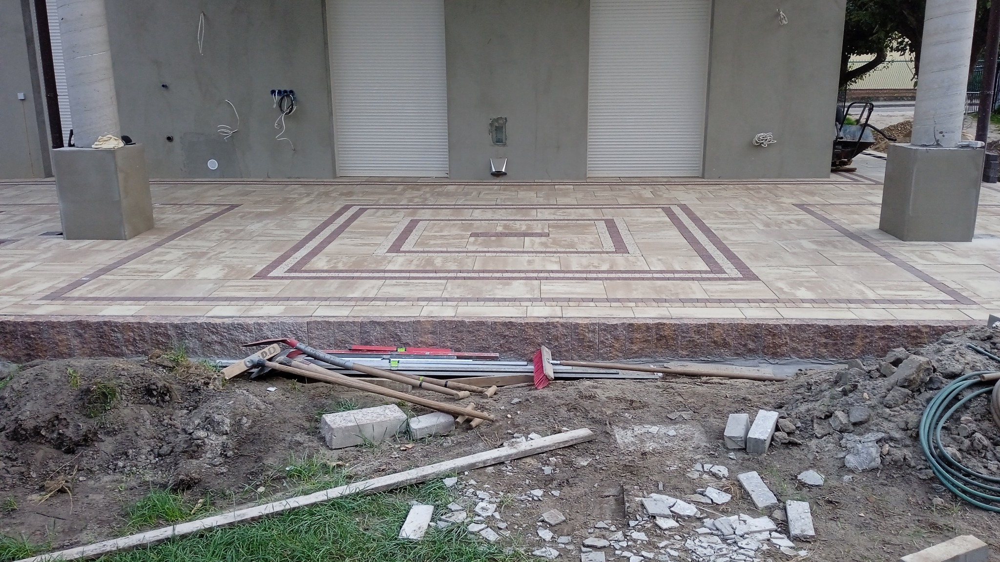 Nowo ułożona, wzorzysta nawierzchnia z kostki brukowej w odcieniach beżu i brązu, otoczona ziemią i narzędziami budowlanymi, z widocznymi elementami elewacji budynku w tle.
