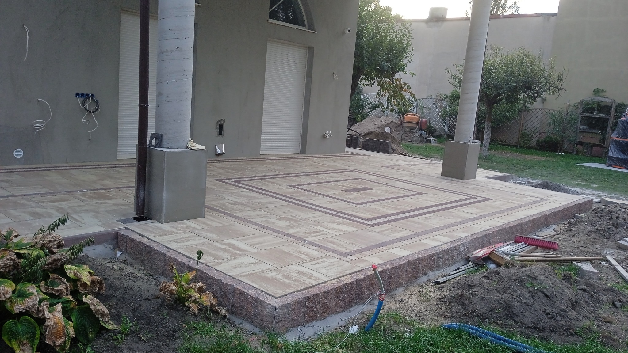 Fragment tarasu z kostki brukowej ułożonej we wzór kwadratu, widoczne narzędzia i resztki materiałów budowlanych na rabacie z ziemią.