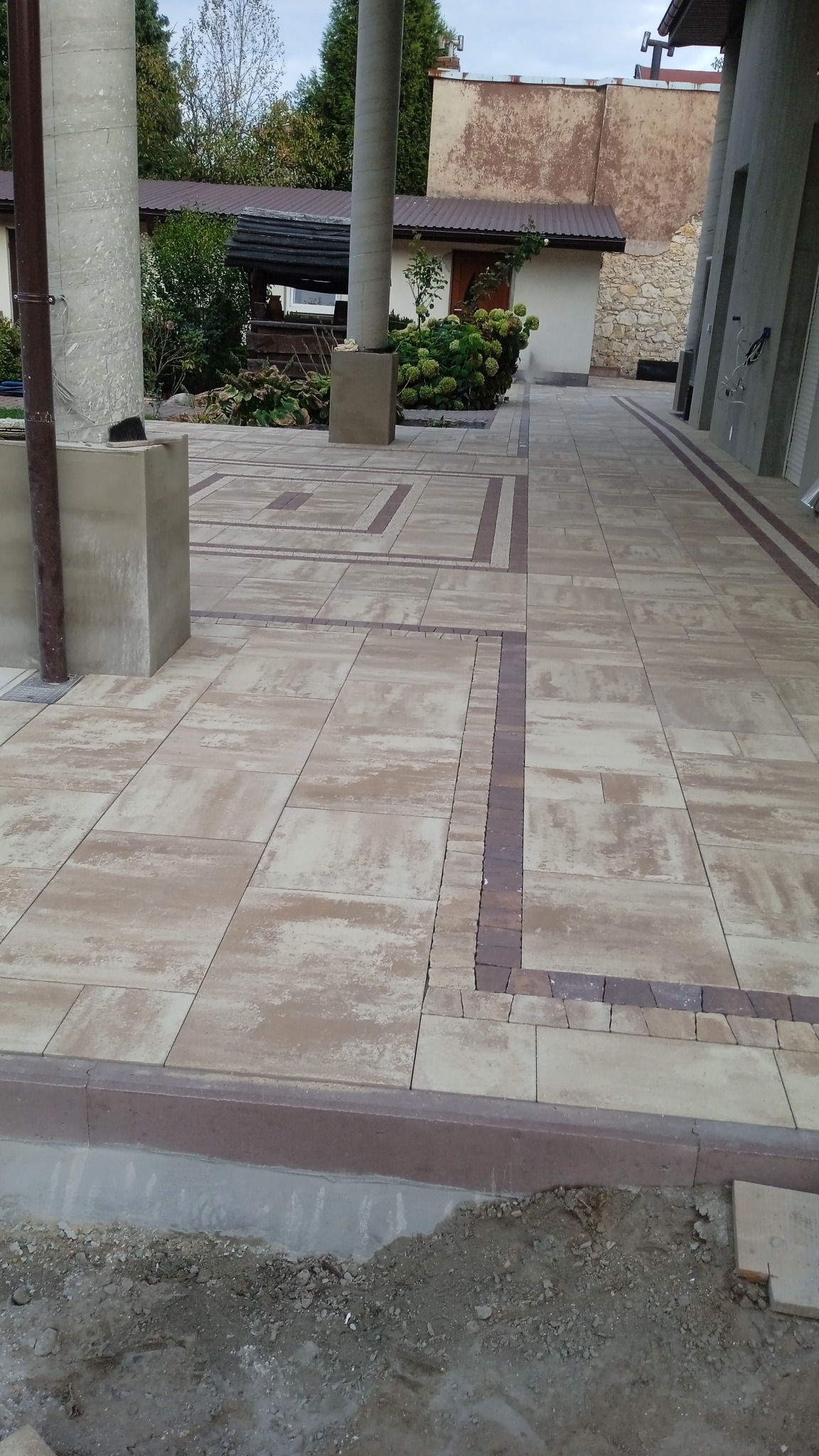 Beżowa kostka brukowa ułożona w geometryczny wzór z bordowymi akcentami, widoczne niedokończone obrzeże i przygotowany grunt pod dalsze prace.