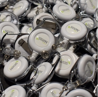 Stos białych identyfikatorów z logo 'SILVAN' i zielonym listkiem, widoczne metalowe klipsy i przezroczyste plastikowe obudowy.