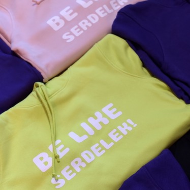 Stos ułożonych bluz z kapturem w kolorach: fioletowym, limonkowym i morelowym. Na każdej bluzie biały nadruk z napisem 'Be Like SERDELEK!'.