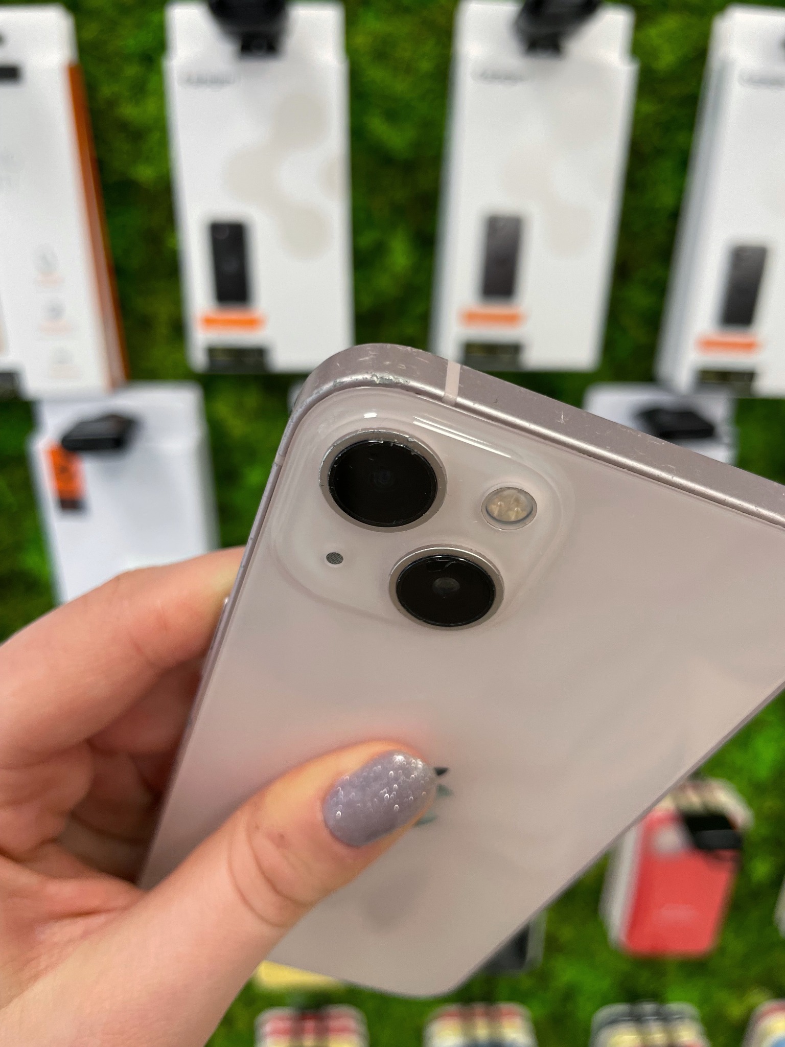 Dłoń trzymająca telefon z widocznym aparatem z dwoma obiektywami i lampą błyskową, w tle pudełka na telefony na sztucznej trawie.