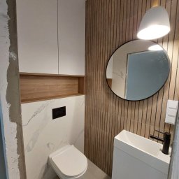 Piotr Guściora Guściora Meble - Nowoczesna łazienka z toaletą podwieszaną, umywalką nablatową, okrągłym lustrem i drewnianą okładziną ścienną z pionowych lameli, oświetlona wiszącą lampą z białym kloszem.