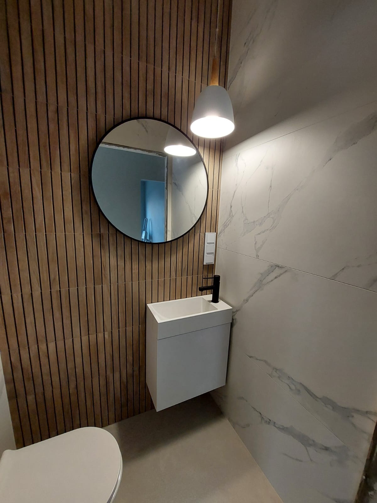 Nowoczesna łazienka z drewnianą ścianą, okrągłym lustrem i wiszącą szafką z umywalką o minimalistycznym designie, oświetlona pojedynczą lampą wiszącą.