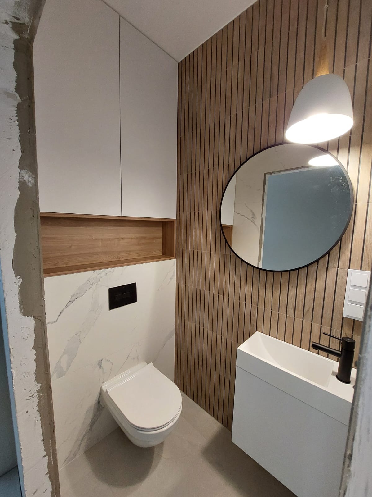 Nowoczesna łazienka z toaletą podwieszaną, umywalką nablatową, okrągłym lustrem i drewnianą okładziną ścienną z pionowych lameli, oświetlona wiszącą lampą z białym kloszem.