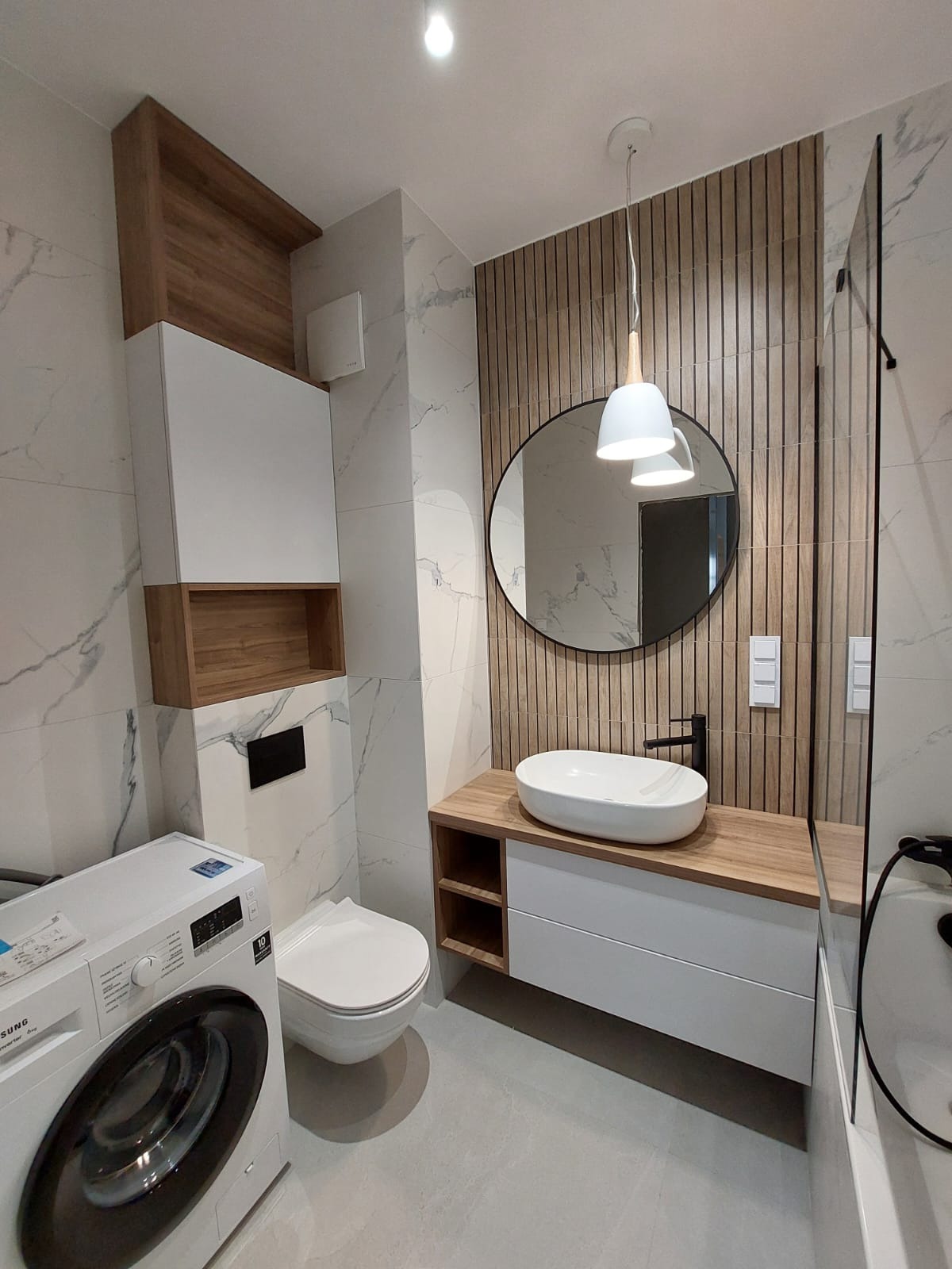 Nowoczesna łazienka z marmurowymi płytkami, drewnianymi lamelami, owalnym lustrem i wiszącą lampą. Uwagę zwraca zabudowa toalety z szafką i pralka po lewej stronie.
